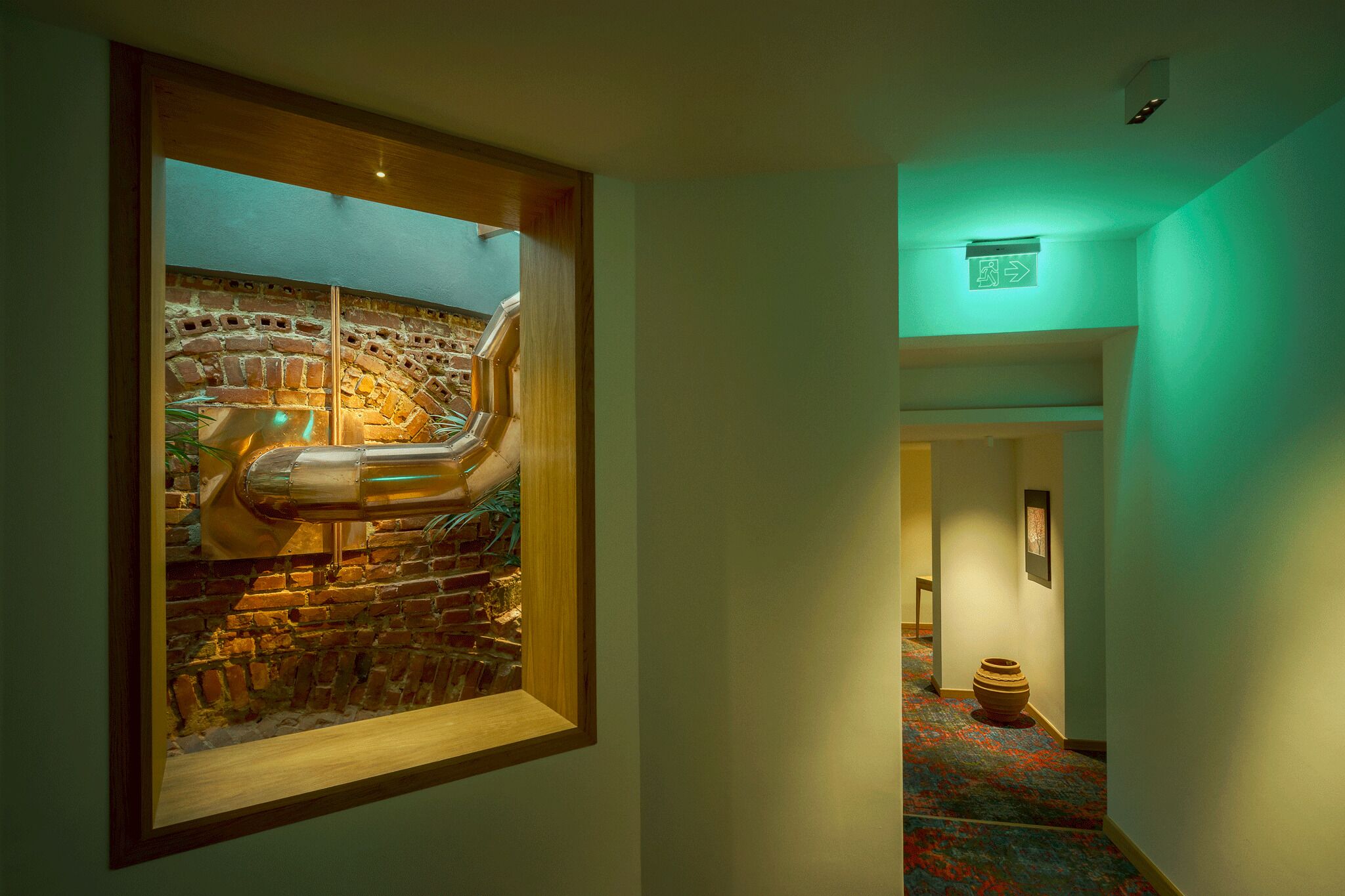 A hallway with a framed picture of a brick wall and a sculpture. There is a green exit sign on the wall and a vase on the floor.