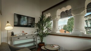 A modern hotel room with a sofa, a small table with a magazine, a lamp, a potted plant, a painting, and a view of buildings through a window with architectural columns. - La Boheme (Bucharest)