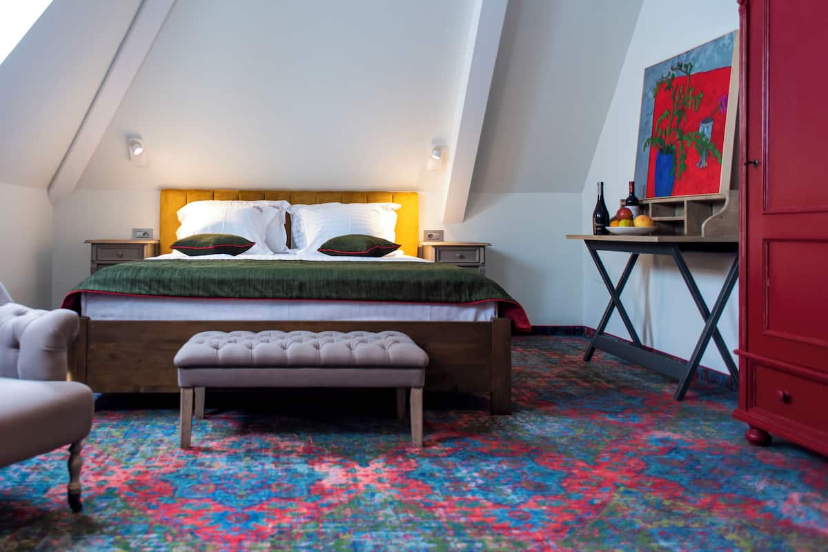 Une chambre avec un plafond en pente, un lit recouvert d’une couverture verte, un fauteuil capitonné, un banc, une petite table avec des bouteilles et un tableau accroché au mur.