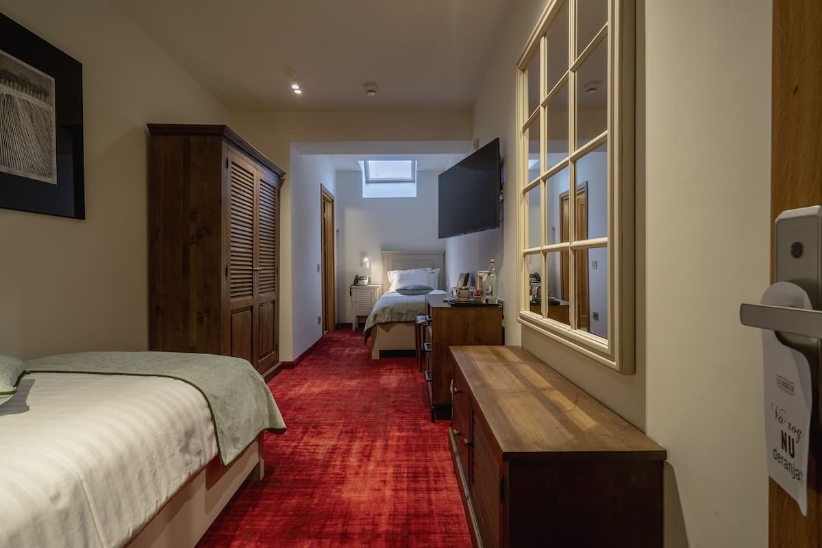 Une chambre d’hôtel étroite, avec un tapis rouge, des meubles en bois, un lit, un bureau et un téléviseur fixé au mur.
