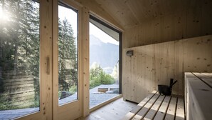 A wooden cabin with large windows, a bench, and a view of a forest and mountains.