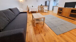 Ein Wohnzimmer mit einer Couch, einem Couchtisch, einem Teppich, einem Esstisch mit Stühlen, einer Küchenzeile und einem Fernseher auf einem Schrank.