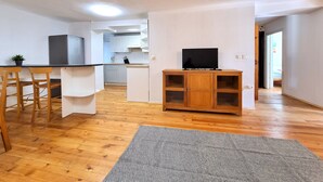 Ein kompakter Wohnbereich mit Küchenzeile, einem Essbereich mit Holzbänken, einem Fernseher auf einem Holzschrank und einem grauen Teppichboden.