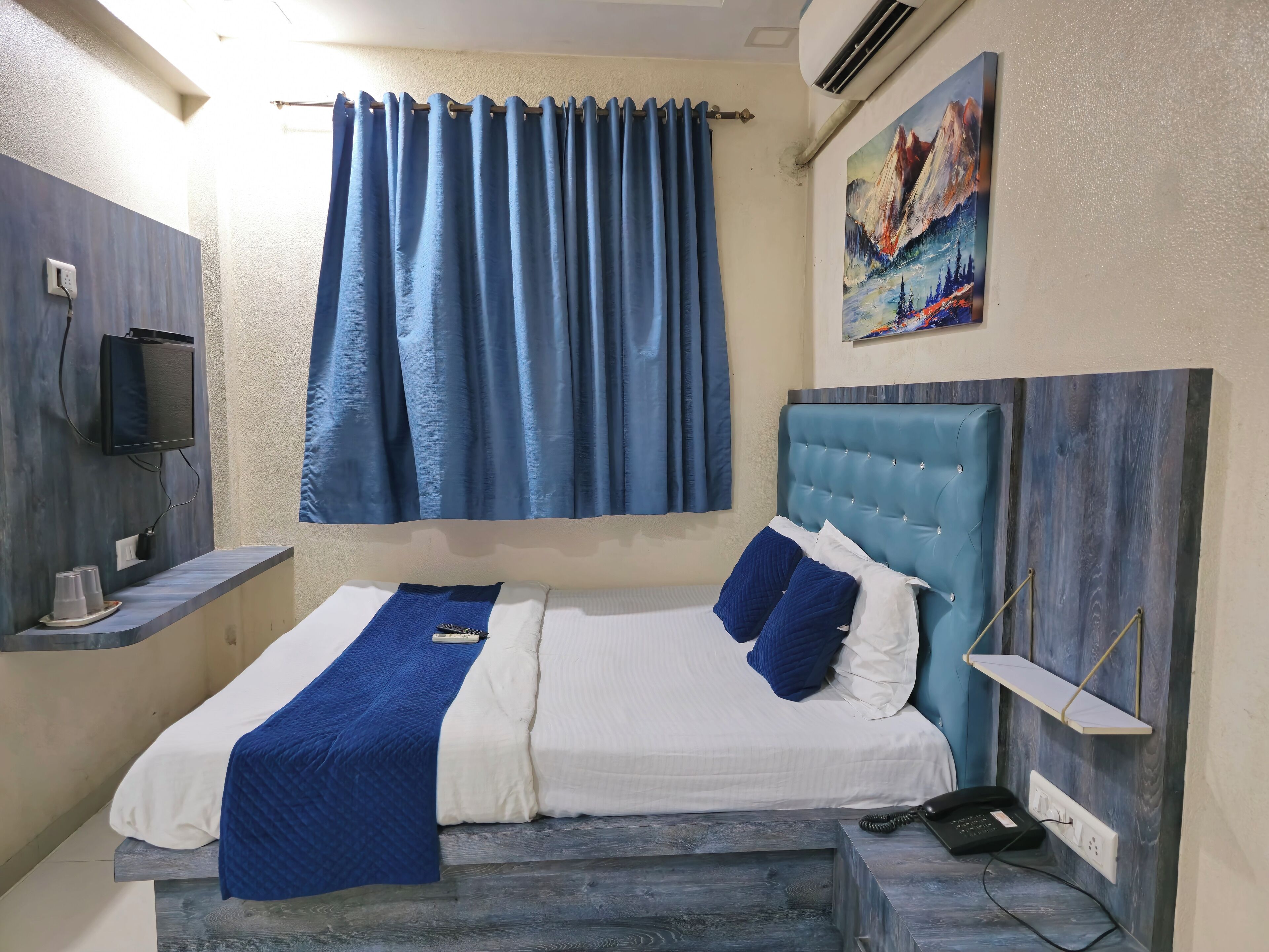 A bedroom with a bed, a TV, a painting, and a blue curtain.