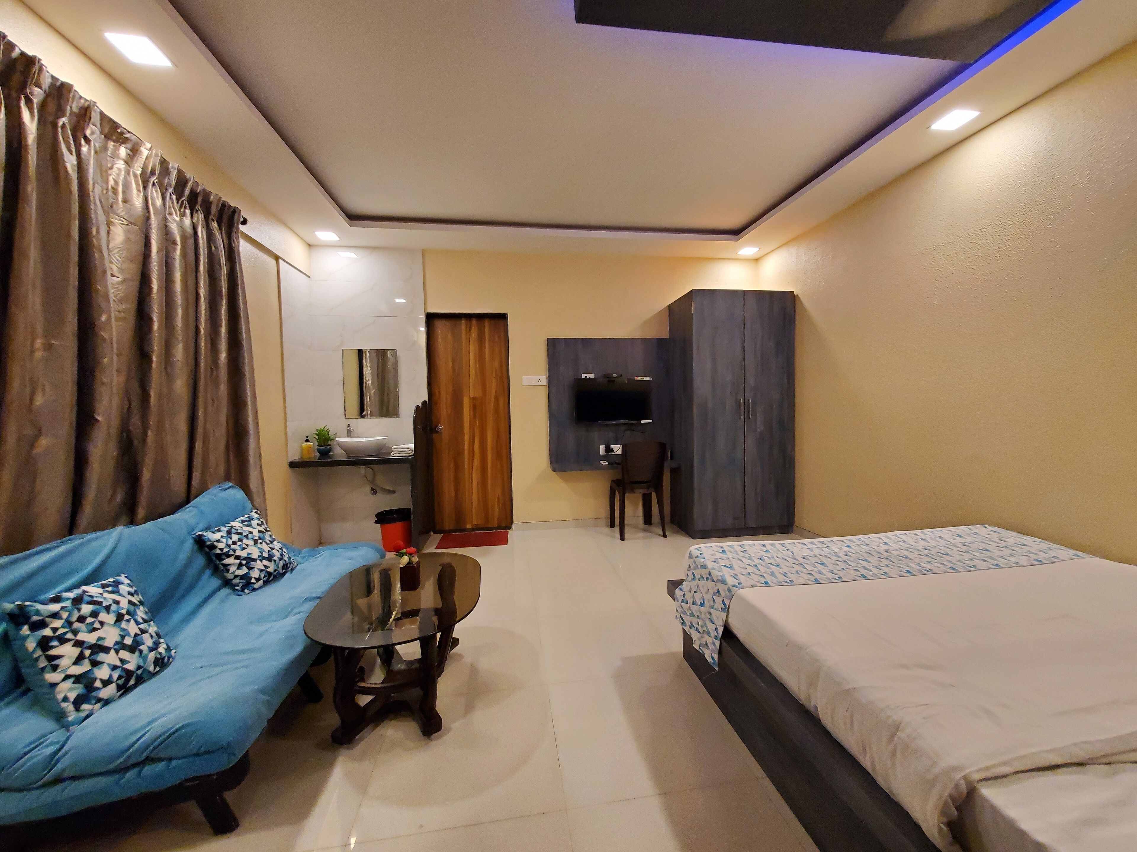A compact hotel room with a blue sofa, a small round table, a bed, a desk with a chair, a wardrobe, and a TV.