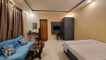 A compact hotel room with a blue sofa, a small round table, a bed, a desk with a chair, a wardrobe, and a TV.