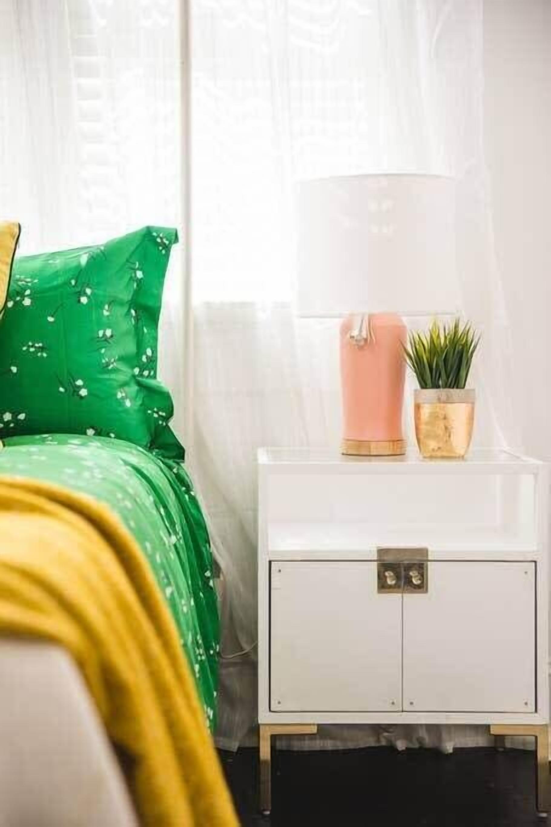A bedroom with a green bedspread, a white nightstand with a pink candle and a small plant.