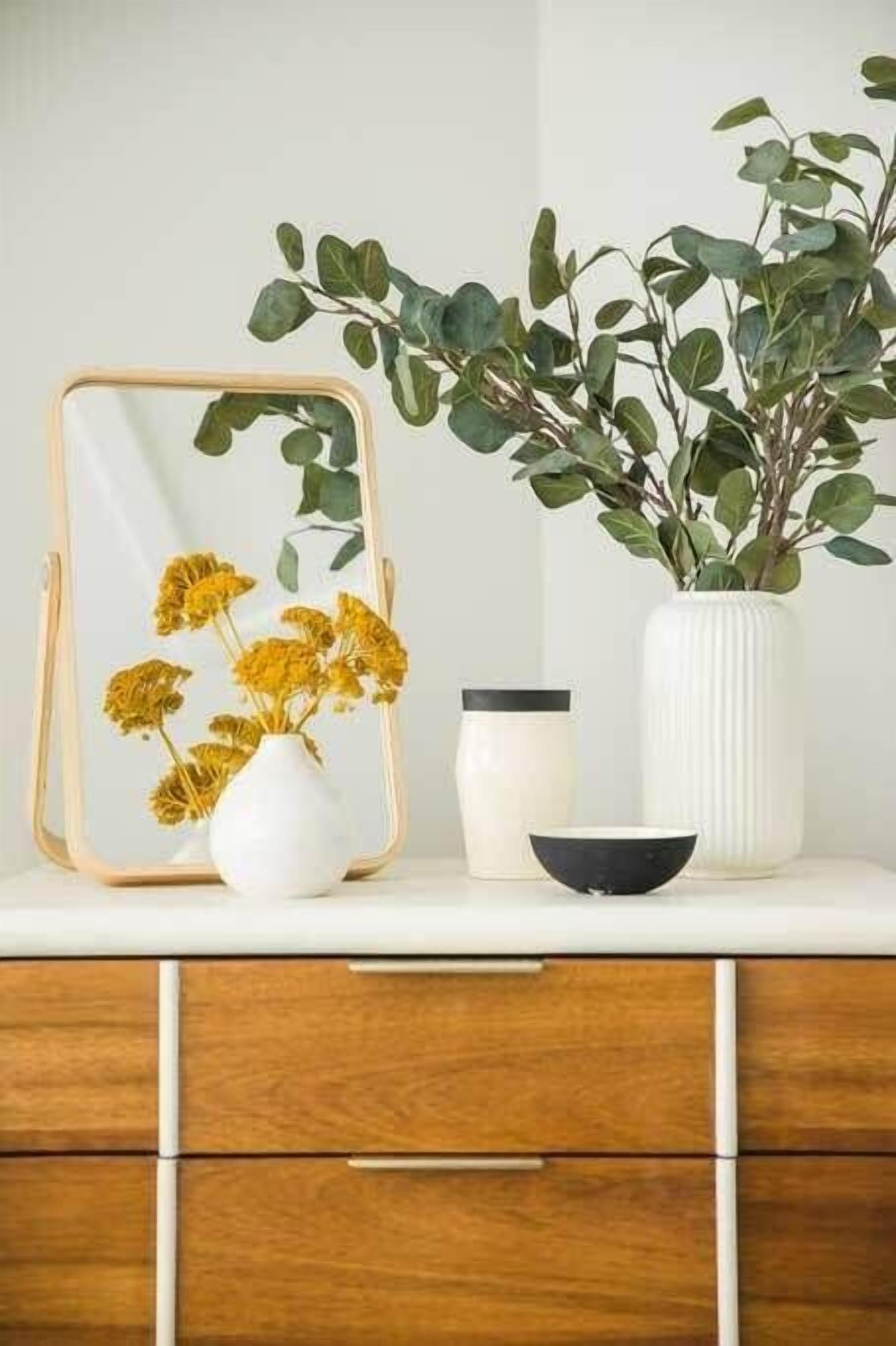 Um penteadeiro de madeira com espelho, um vaso branco com flores amarelas, um vaso branco com folhas verdes e uma tigela preta.