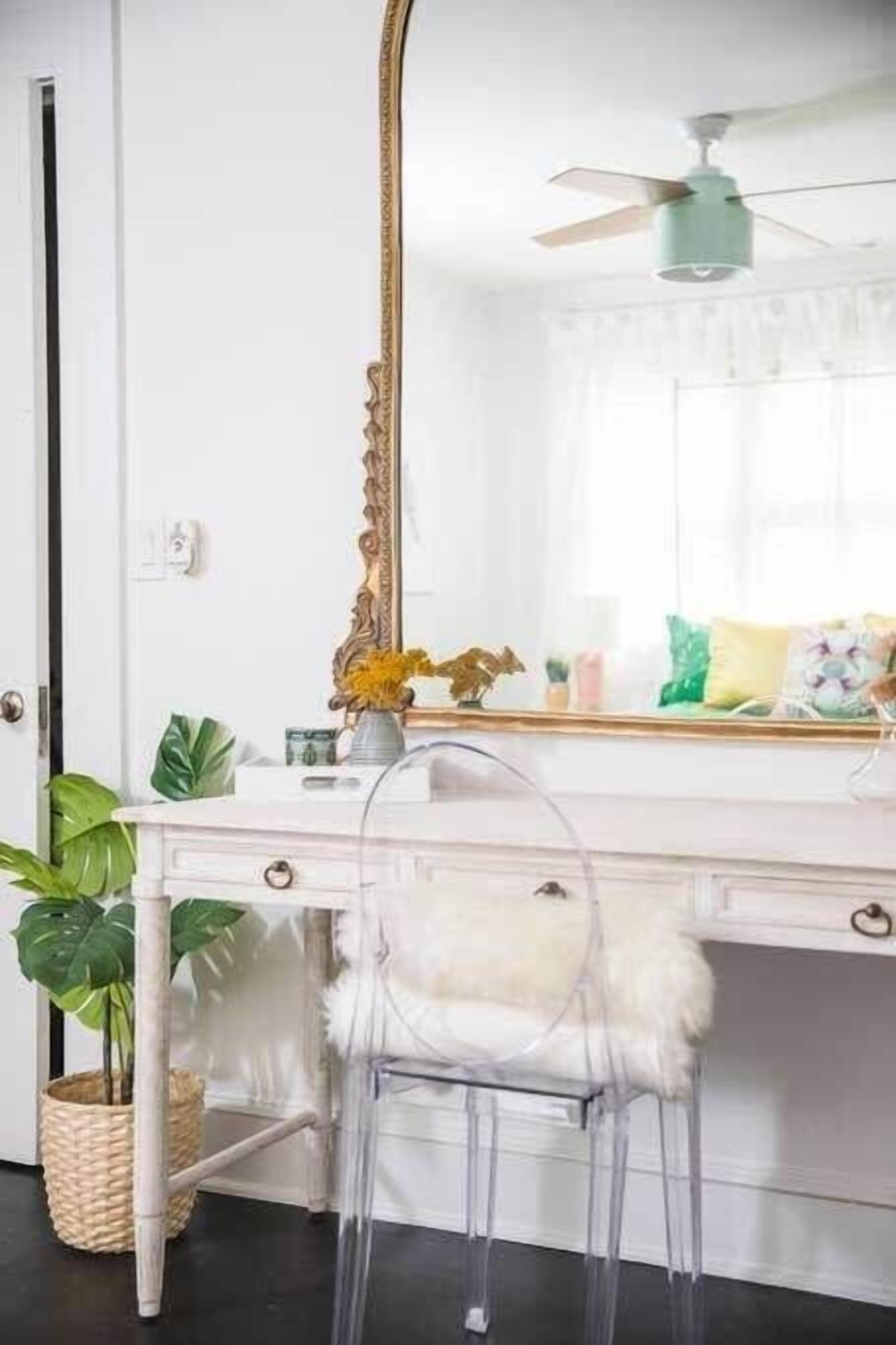 Un escritorio blanco con un espejo grande, un ventilador de techo y una silla transparente.