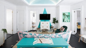 A modern living room with a teal sofa, a coffee table with a notebook, a TV mounted on a teal wall, and a green plant.
