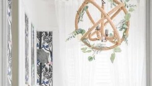 A room with a woven chandelier, a wicker chair, and a white wall with artwork.