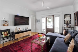 A living room with a black sofa, a red patterned rug, a glass coffee table, a black chair, a TV on the wall, and a door with a wreath.