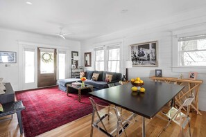 A spacious living room with a dark sofa, a red rug, a dining table with chairs, and a large window with a view of trees.
