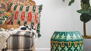 A room with a wooden bed, patterned pillows, a green decorative stool, and a large plant.