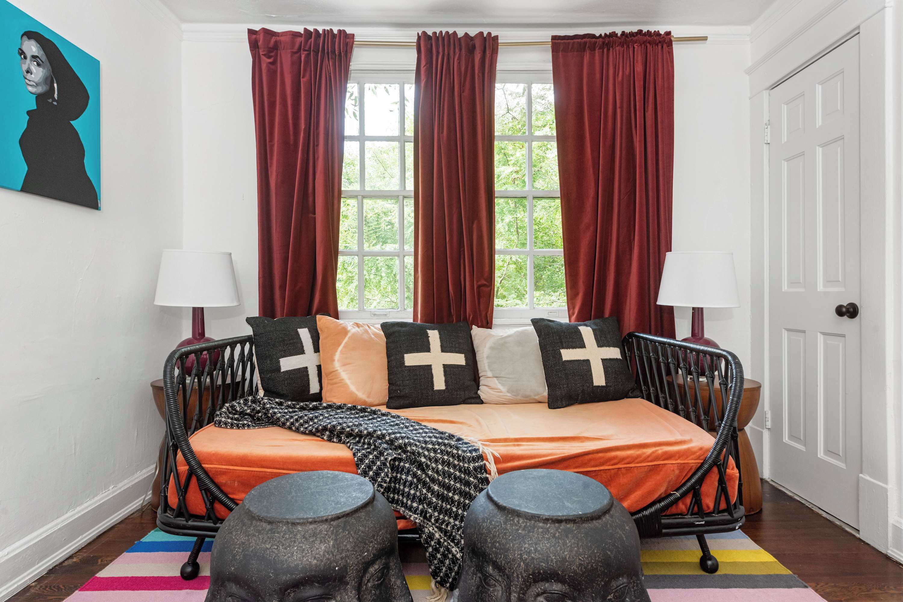 A bedroom with a bed, two bedside lamps, a window with curtains, a door, and a colorful rug.