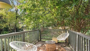 A wooden deck with two white wire chairs and a round woven table, surrounded by lush greenery. - Lucky Leopard (Atlanta)