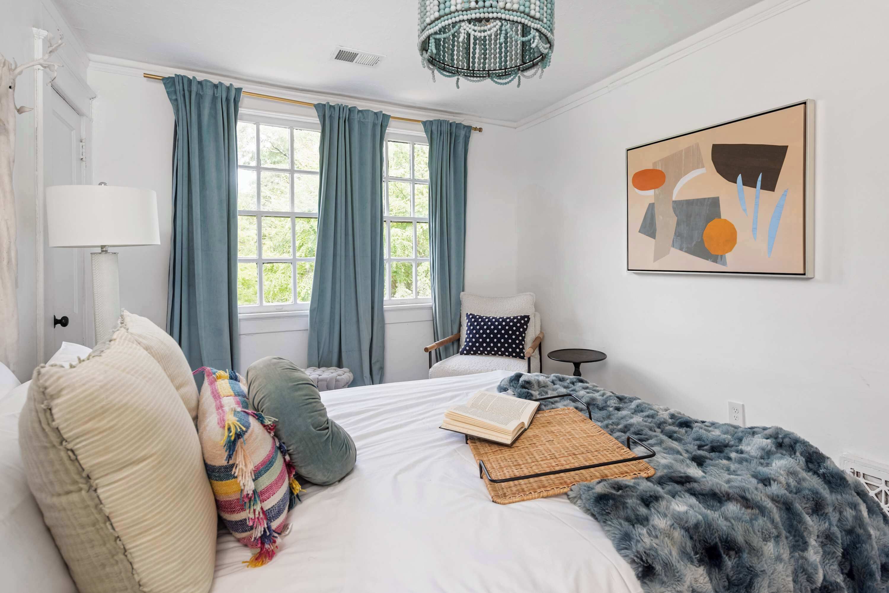 A bedroom with a bed, a chair, a lamp, a window with curtains, and a painting on the wall.