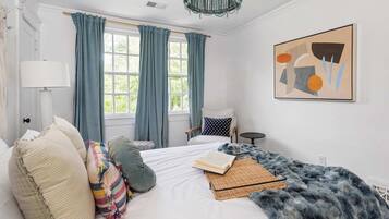 A bedroom with a bed, a chair, a lamp, a window with curtains, and a painting on the wall.