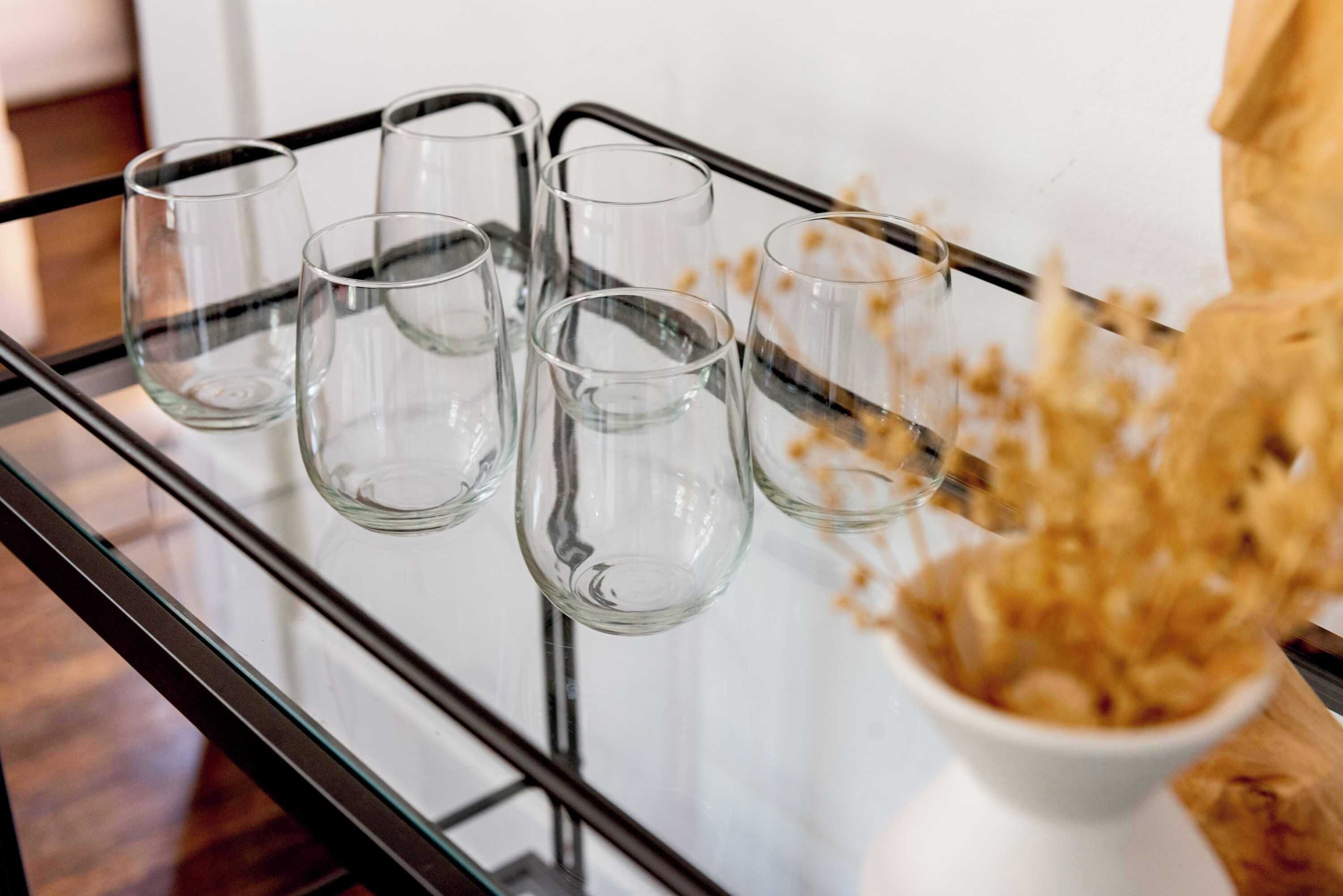 Une table en verre, avec six verres transparents et un vase blanc contenant des fleurs séchées.
