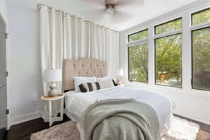 A bedroom with a tufted headboard, white bedding, a fluffy rug, and a view of trees through large windows.