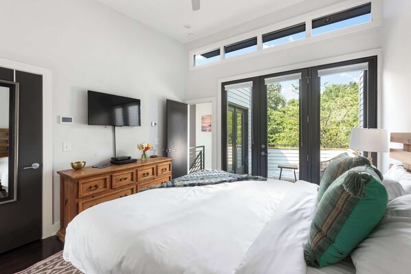 A bedroom with a large bed, a TV mounted on the wall, a wooden dresser, and a view of the outdoors through a sliding door.