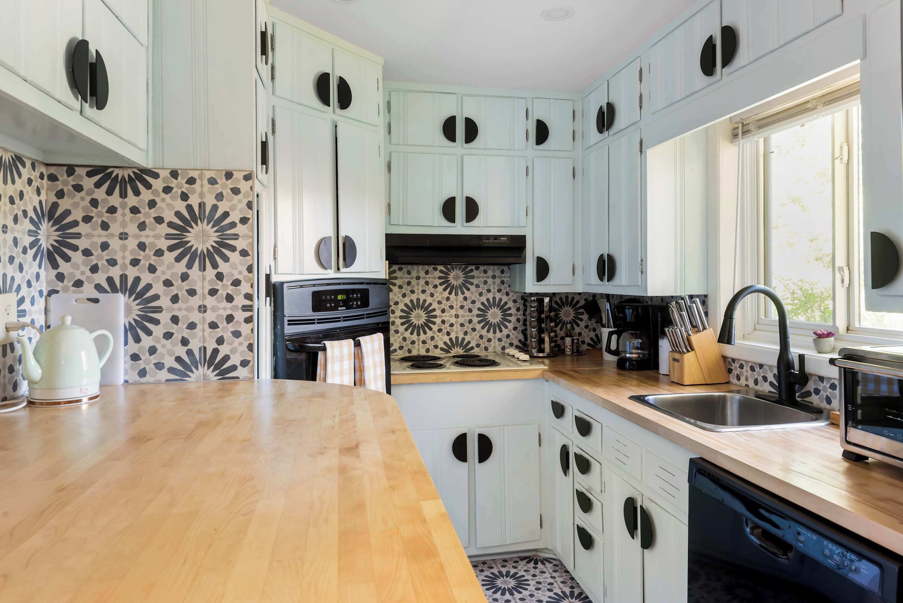 A kitchen with light blue cabinets, a wooden countertop, a stove, a sink, and a window.