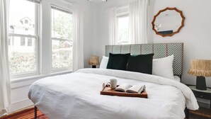 A neatly made bed with a tray of books and a cup, large windows with curtains, a mirror, and a patterned headboard. - Morehaus (Atlanta)