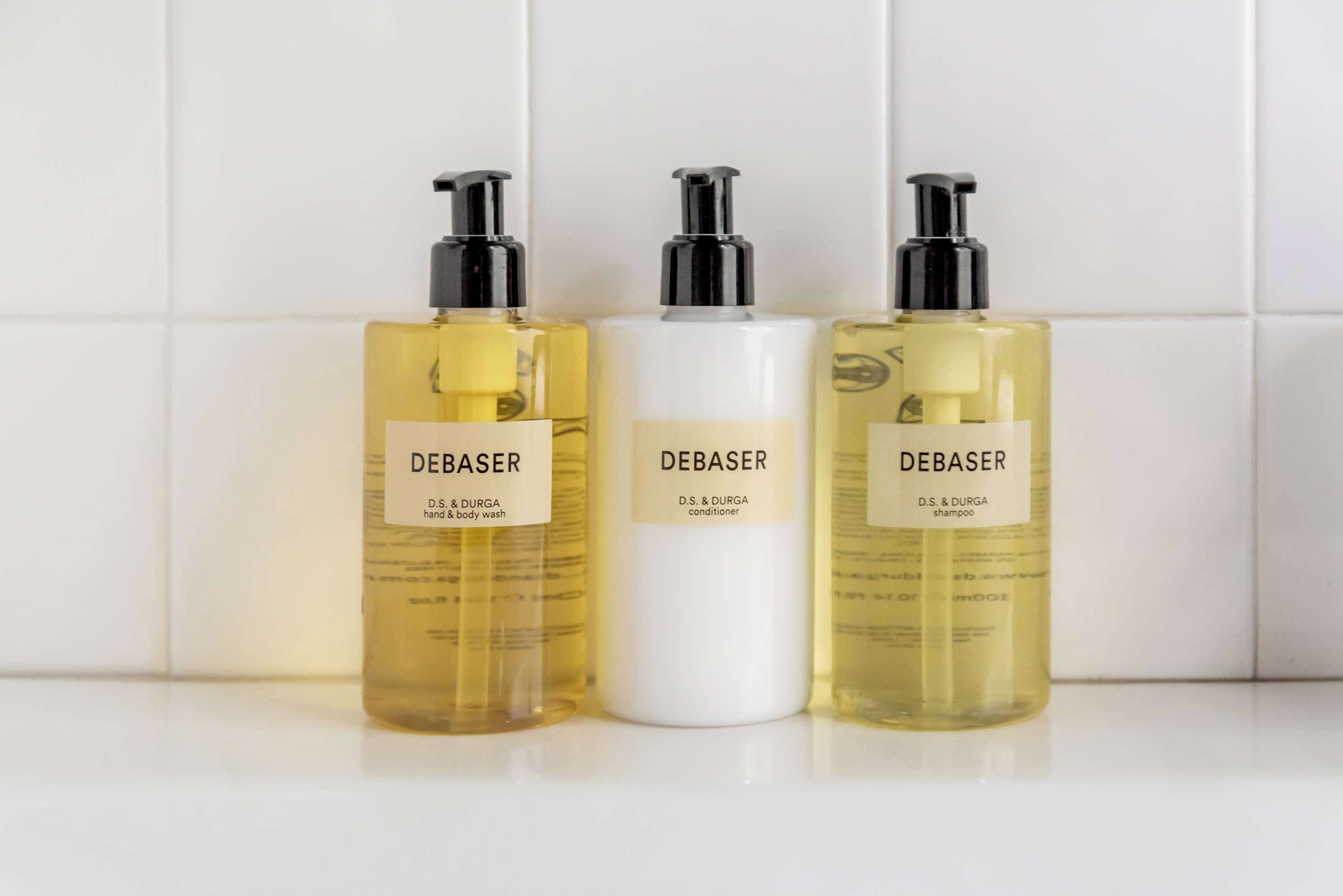 Tres botellas de productos Debaser para cuerpo y cabello: lavado corporal, acondicionador y champú, sobre una superficie de azulejos blancos.