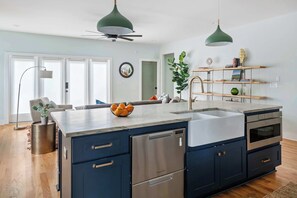 Una cocina moderna con una isla grande, electrodomésticos de acero inoxidable y un horno empotrado. Dos lámparas colgantes iluminan la isla, y hay una planta sobre la encimera.