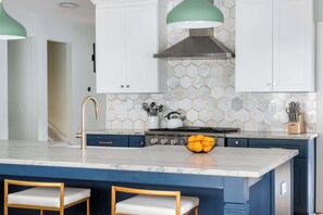Una cocina moderna con encimera de mármol, una isla central con taburetes, armarios blancos y un salpicadero hexagonal.
