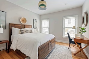 Un dormitorio con cama de madera, ropa de cama blanca, un adorno circular en la pared, un escritorio con silla y una ventana con persianas.