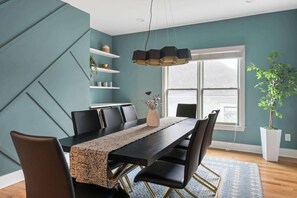A dining area with a long table, chairs, a hanging light fixture, and a potted plant.