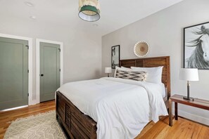 A bedroom with a wooden bed, white bedding, a woven rug, a bedside table with a lamp, and a wall-mounted artwork.