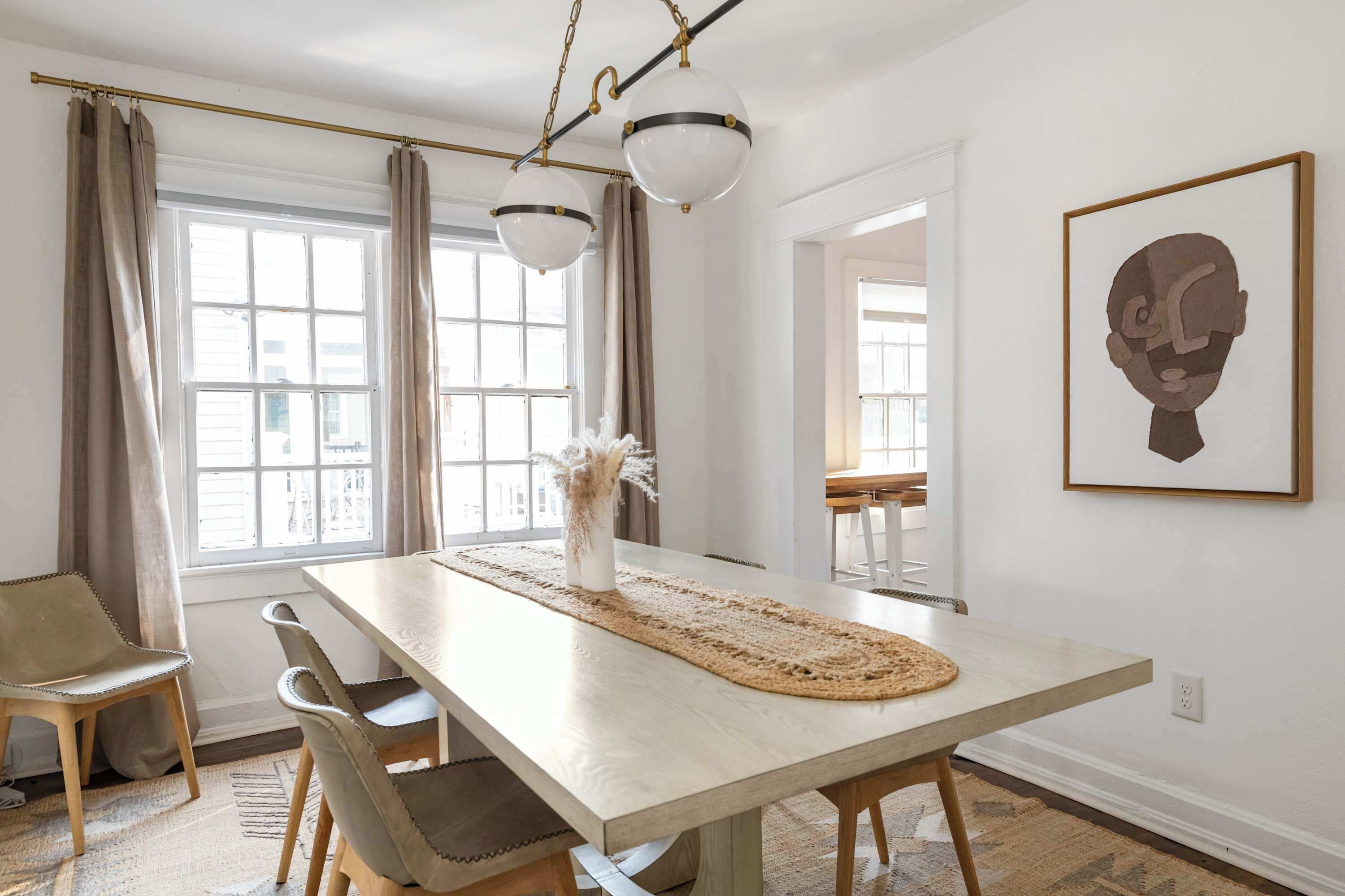 A dining area with a long table, chairs, a vase with dried flowers, and a framed artwork on the wall.