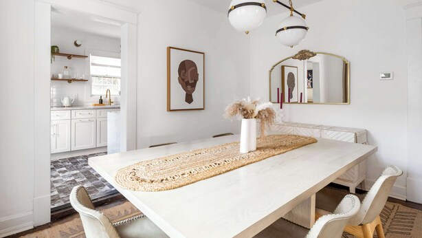 A dining area with a long table, white chairs, a woven placemat, and a vase with dried flowers. There is a kitchen visible in the background with white cabinets and a window. - Golden Gazelle (Atlanta)