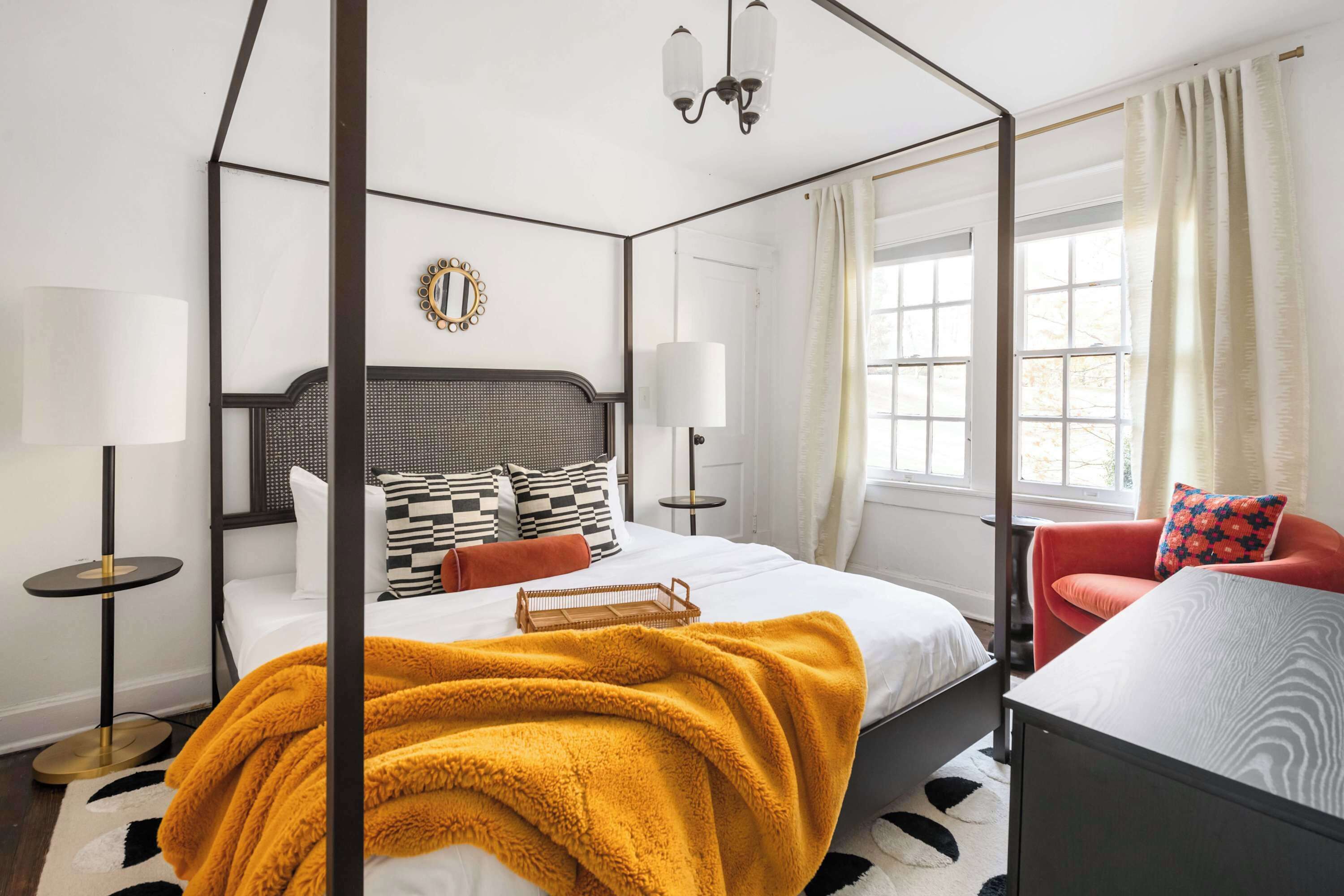 A four-poster bed with a mustard-colored blanket, a mirror on the wall, and a red chair.