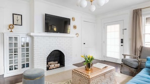 A living room with a white brick fireplace, a flat-screen TV mounted above it, a blue sofa, a patterned ottoman, and a wooden coffee table. - Golden Gazelle (Atlanta)
