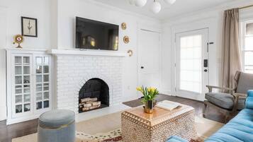 A living room with a white brick fireplace, a flat-screen TV mounted above it, a blue sofa, a patterned ottoman, and a wooden coffee table.