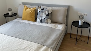 A neatly made bed with a grey and white duvet, two pillows (one yellow and one with a geometric pattern), and a grey headboard. There are two bedside tables with lamps and a wall socket. - Terrace Flair (Wien)