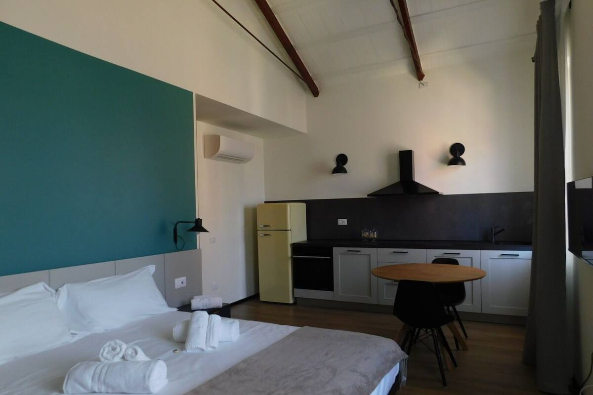 A hotel room with a bed, a small kitchenette, a dining table, and a ceiling with exposed beams.