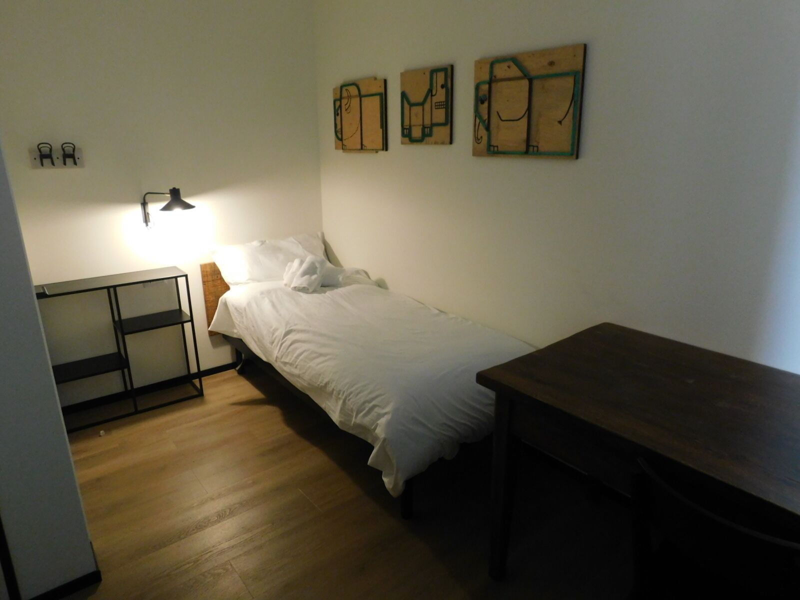 A small, sparsely furnished room with a single bed, a desk, and a table. There are three wall-mounted artworks above the bed.