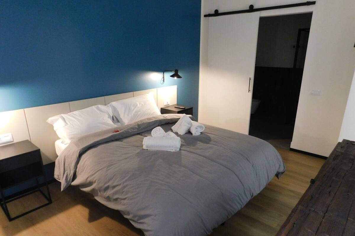 A neatly made bed with a grey blanket, white pillows, and neatly folded towels. There is a bedside table with a lamp, a wooden bench, and a door leading to another room.
