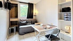 Una habitación pequeña con sofá, una mesita, una silla, televisor montado en la pared y ventana con cortinas.