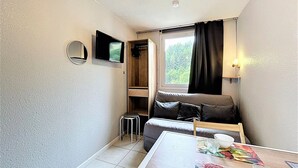Una habitación pequeña con cama, una mesita, una silla, televisión y ventana con cortinas.