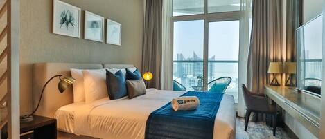 A hotel room with a large bed, a desk with a chair, a view of the city through large windows, and artwork on the wall.
