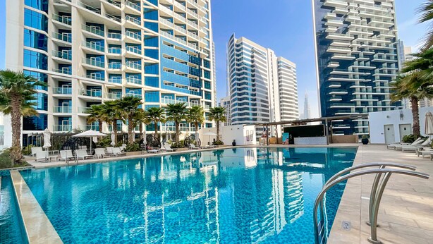 A swimming pool with lounge chairs and umbrellas, surrounded by tall buildings with balconies. - HiGuests - DAMAC Maison Prive Tower A (Dubai)