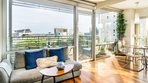 A modern living room with a sofa, a coffee table, and a dining area with chairs. Large windows provide a view of the cityscape.