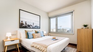 A neatly arranged bedroom with a bed, bedside table, lamp, and a window with a view of the city.