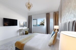 A modern hotel room with a large bed, a flat-screen TV mounted on the wall, a desk, and a view of the sea through the window.