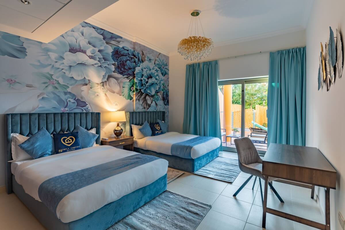 A hotel room with two beds, a desk, a chair, a large floral mural, and a view of the outdoors.